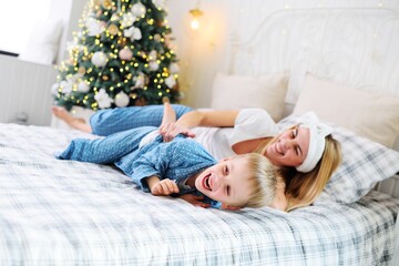 Obraz na płótnie Canvas young mother laughs and tickles her young son in pajamas while lying on the bed against the background of a Christmas tree.