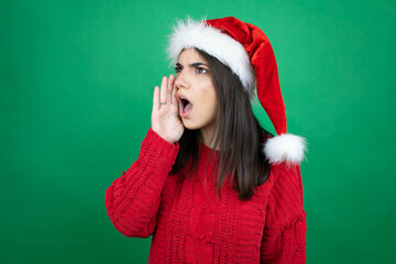 Young beautiful woman wearing Christmas Santa hat over isolated green background shouting and screaming loud to side with hand on mouth