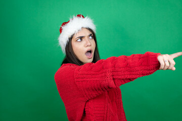 Young beautiful woman wearing Christmas Santa hat over isolated green background pointing with finger surprised ahead, open mouth amazed expression, something on the front