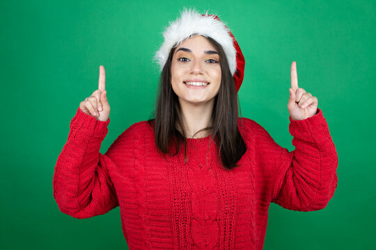 Young beautiful woman wearing Christmas Santa hat over isolated green background smiling, looking at the camera and pointing up with fingers and raised arms