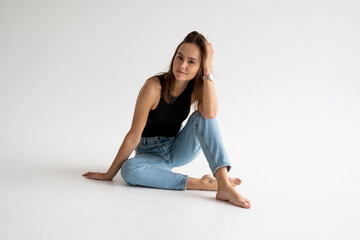 Fototapeta premium Portrait of happy young pensive woman posing in black underwear and blue jeans without a shoes, sitting on a white floor in white studio. Model tests of pretty girl in basic clothes on cyclorama.