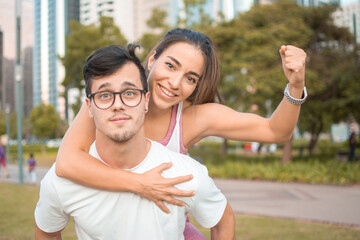 Happy sporty couple piggy back. Handsome young man wearing eyeglasses carry on his back young woman flexing muscle with fist up outdoors.
