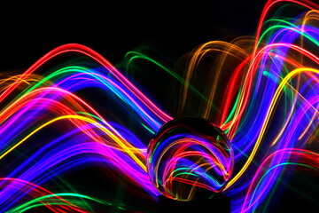 Long exposure photograph of neon multi colour in an abstract swirl, parallel lines pattern against a black background with reflections in a glass crystal ball. Light painting photography.