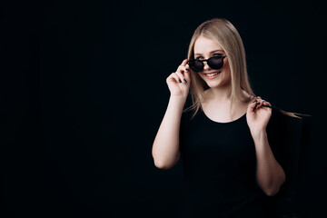 Attractive woman in trendy sunglasses holding black shopping bags while standing over black studio background. Happy blonde enjoying shopping time.