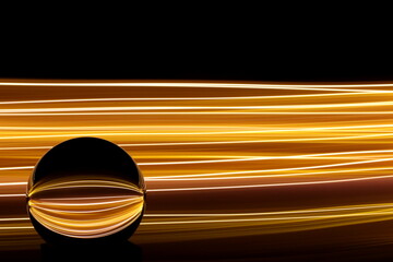 Long exposure photograph of neon gold colour in an abstract swirl, parallel lines pattern against a black background with reflections in a glass crystal ball. Light painting photography.