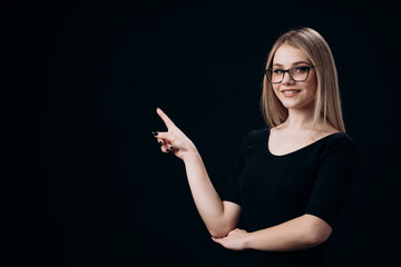 Adorable young woman in eyeglasses standing over black background and pointing on empty space with forefinger. Pretty lady with blond hair presenting something.