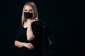 Obraz premium Pretty young lady in medical protective mask holding paper bags over black studio background. Concept of sales and purchase.