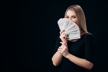 Happy young blonde covering half of face with fan of dollar banknotes. Isolated over black studio background. Concept of rich and luxury lifestyle.