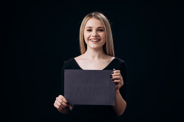 Fototapeta premium Portrait of smiling young woman standing over black studio background and holding board with empty space. Concept of black friday and sales.