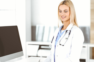 Blond female physician is standing at her workplace near desktop computer. Woman-doctor is excited and happy of her profession. Medicine concept