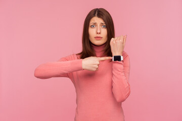 Displeased impatient woman with brown hair in pink sweater pointing finger on her wrist clock...