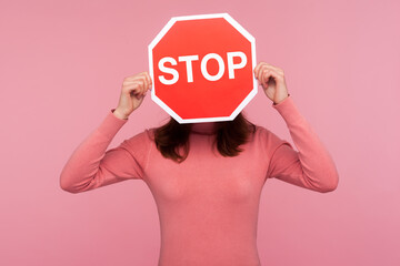 Brunette woman in pink sweater hiding face behind stop sign, way prohibited, stop violence. Indoor studio shot isolated on pink background