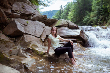 Young Woman in black leggings is sitting at stone near at Splashing Waterfall. Peaceful Caucasian Traveler Sit at Beautiful Stream.