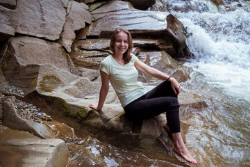 Young Woman in black leggings is sitting at stone near at Waterfall. Peaceful Caucasian Traveler Sit at Beautiful Stream.
