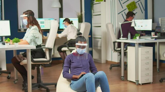 African employee with protection mask against covid-19 pandemic sitting on armchair in middle of office room holding digital tablet. Multiethnic business team working respecting social distance