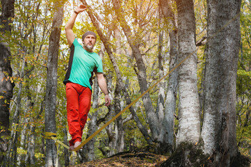 Naklejka na ściany i meble A bearded man in age balances while sitting on a taut slackline in the autumn forest. Outdoor Leisure