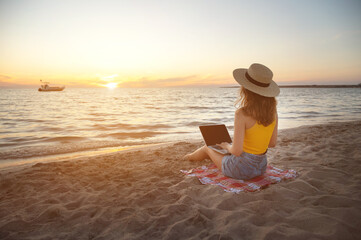 Young woman freelancer in denim shorts and a straw hat with a laptop on her legs sits on the sandy seashore at sunset. Remote work via the Internet in a warm effort by the ocean