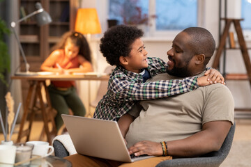 Happy affectionate African boy embracing his father with laptop against daughter