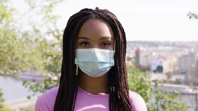 A young black woman in a face mask looks at the camera - face closeup - trees and a cityscape in the blurry background