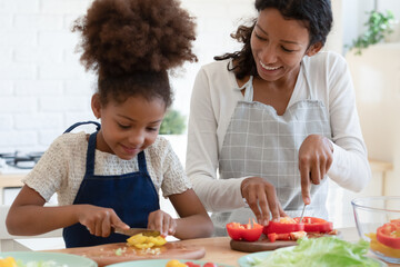 Fototapeta na wymiar Culinary is easy. Inspired happy african american mother and tween daughter prepare vegan food, grown elder and small younger sisters trying new cooking recipe, cutting vegetables talking at kitchen