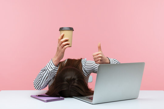 I am fine. Extremely tired brunette woman lying on desk at workplace holding paper cup of coffee and showing thumbs up, lack of energy, procrastination. Indoor studio shot isolated on pink background