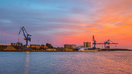 Massive blue cranes unload cargo in a seaportduring sunset