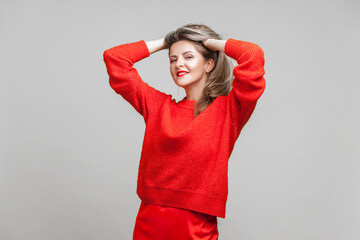 Portrait of impressive happy blonde woman with red lipstick in bright sweater standing with hands in hair, looking at camera with seductive smile. indoor studio shot isolated on gray background