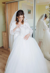 Naklejka premium Beautiful happy young bride in a beautiful robe, peignoir, standing by the mirror holding a wedding dress in her hands.