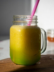 sunlit glass jar with yellow smoothies. There's a drinking tube in the jar. Wooden background. Vertical photo