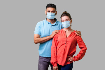 Portrait of happy young couple with surgical medical mask standing together, looking at camera with toothy smile, caring man holding female shoulders. isolated on gray background, indoor studio shot