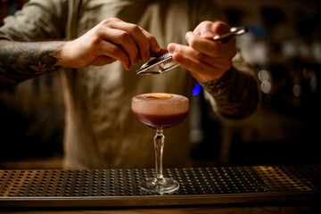 male bartender grates cinnamon over glass of cocktail with foam