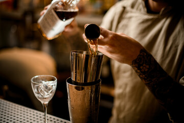male barman pours alcoholic drink from metal jigger into shaker on bar counter