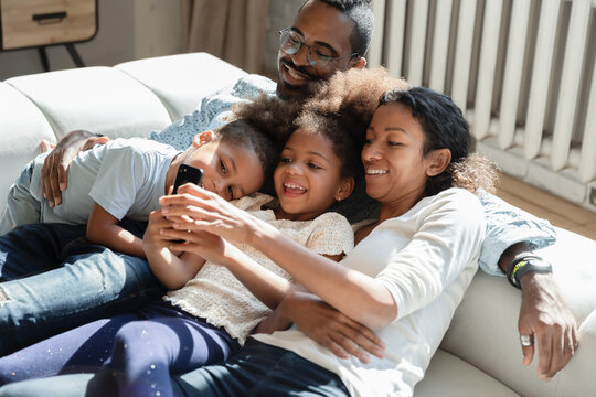 Family picture on phone. Happy loving african family couple with tween daughter and little son preschooler cuddling on couch at home taking selfie on cellphone, making video call, watching cute movie