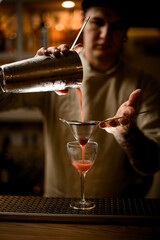 young male bartender holds sieve over wine glass and gently pours cocktail from shaker.