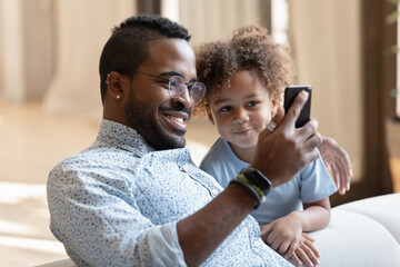 Say cheese, sonny. Happy black dad sitting on couch in living room cuddling cute little boy and taking selfie on smartphone, smiling african father showing son kid funny video or cartoon on cellphone