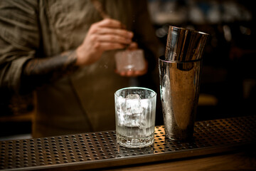 transparent old fashioned glass with ice and shaker stand on bar with bartender in background