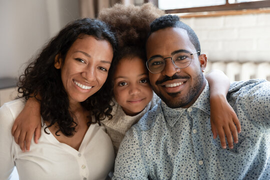 Mom Dad And Me. Cute Adorable Black Child Preteen Girl Adopted Daughter Posing On Portrait Between Happy Affectionate African Foster Mother And Father Holding Arms On Their Shoulders Looking At Camera