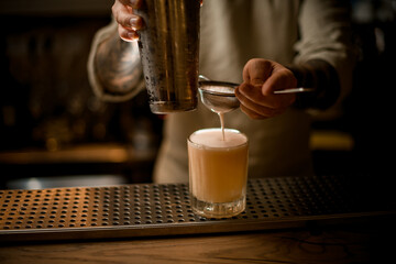 male bartender filters whipped cocktail through sieve into old fashioned glass on bar counter