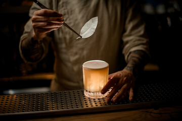 Fototapeta na wymiar barman holds tweezers with decorative leaf over old fashioned glass of alcoholic cocktail
