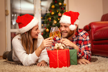 Obraz na płótnie Canvas Beautiful young couple in love spending Christmas day together at home, having lying near nicely decorated Christmas tree, drinking champagne and having fun
