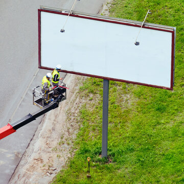 Two Men In A Lift Basket Change Image On A Billboard Near A Road