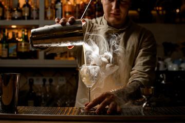 male barman pours steaming drink from steel shaker cup into wine glass with ice cubes.