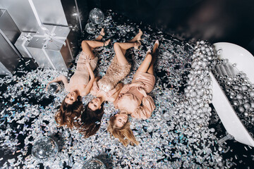 Three beautiful woman's in an evening dress lies on a shiny confetti on a black background with a disco ball. New year 2021