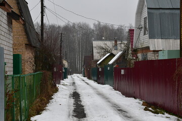 Street in a horticultural Association in late autumn. High metal fences. There is snow on the road. On the horizon is a bare birch forest. Country houses behind high fences.