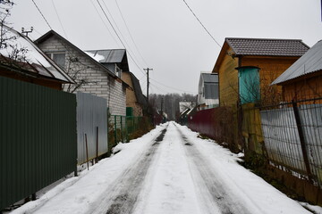 Fototapeta na wymiar Street in a horticultural Association in late autumn. High metal fences. There is snow on the road. On the horizon is a bare birch forest. Country houses behind high fences.