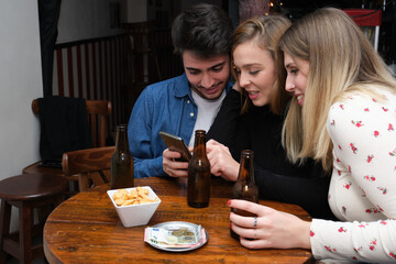 Three friends seeing something on their smartphone, with drink, snacks and money on the table. Bar routine concept.