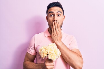 Young handsome romantic man with beard holding bouquet of flowers over pink background covering...