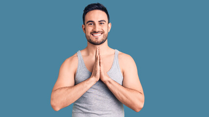 Young handsome man wearing swimwear and sleeveless t-shirt praying with hands together asking for forgiveness smiling confident.