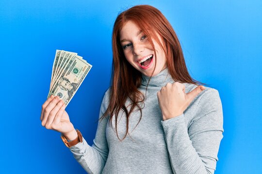 Young red head girl holding 20 dollars banknotes pointing thumb up to the side smiling happy with open mouth