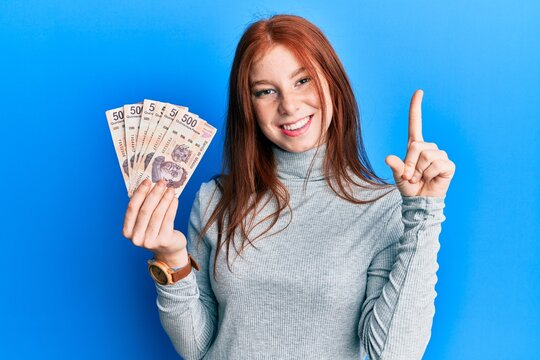 Young red head girl holding 500 mexican pesos banknotes smiling with an idea or question pointing finger with happy face, number one